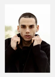 Close-up portrait of a young man with a buzz cut holding the collar of his black coat against a bright, hazy outdoor background.