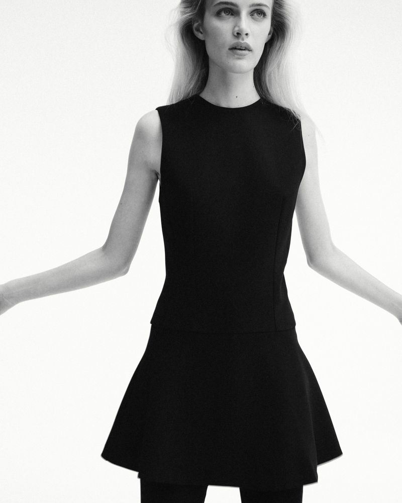 A woman wearing a sleeveless black top and flared mini skirt posing against a bright white seamless background in black and white.