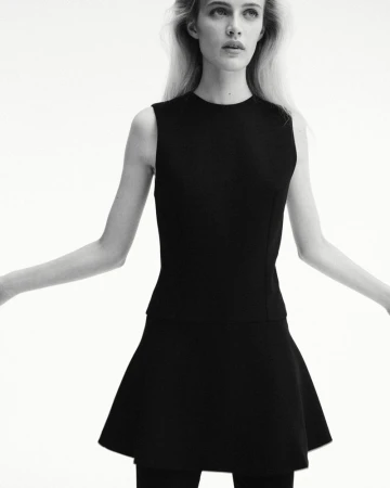 A woman wearing a sleeveless black top and flared mini skirt posing against a bright white seamless background in black and white.