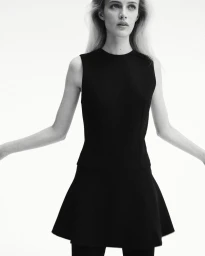 Fashion model in a structured sleeveless black mini dress with flared hem posing against a pure white seamless background.