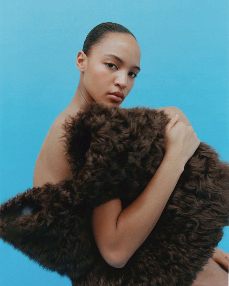 Model Laura Savy holding a textured dark brown shearling garment against a vibrant blue studio backdrop.