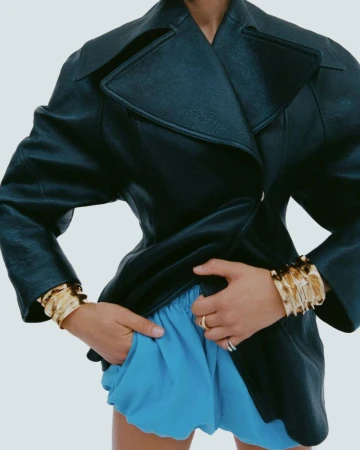 Model wearing a structured dark leather jacket, bright blue shorts, and chunky gold cuffs against a pale studio backdrop.