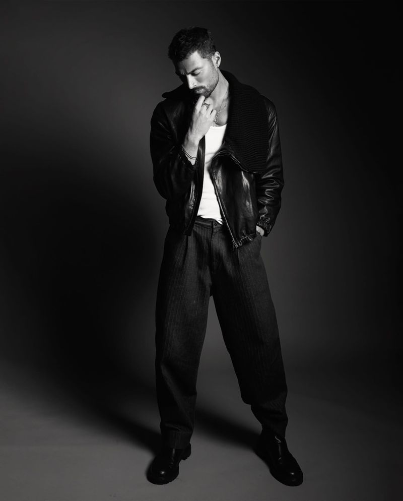 Full-length black and white portrait of Simone Susinna wearing an open black leather jacket, white tank top, and wide-leg trousers.