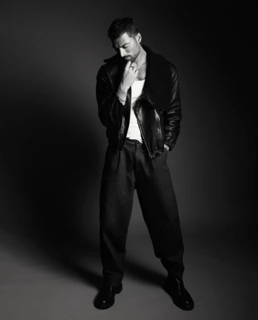 Full-length black and white portrait of Simone Susinna wearing an open black leather jacket, white tank top, and wide-leg trousers.