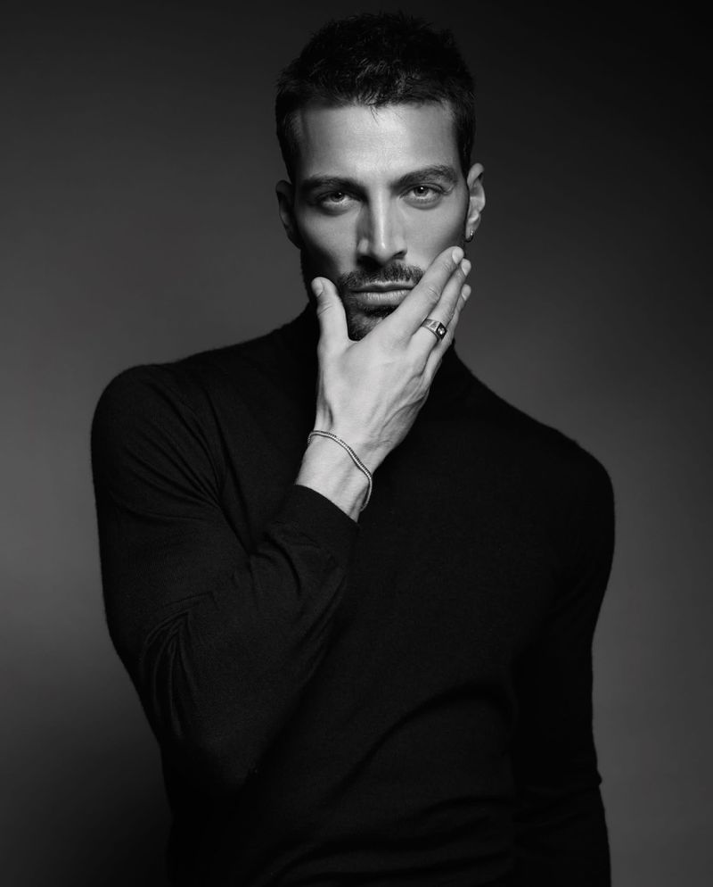 Black and white portrait of Simone Susinna wearing a black turtleneck with his hand touching his chin against a dark backdrop.