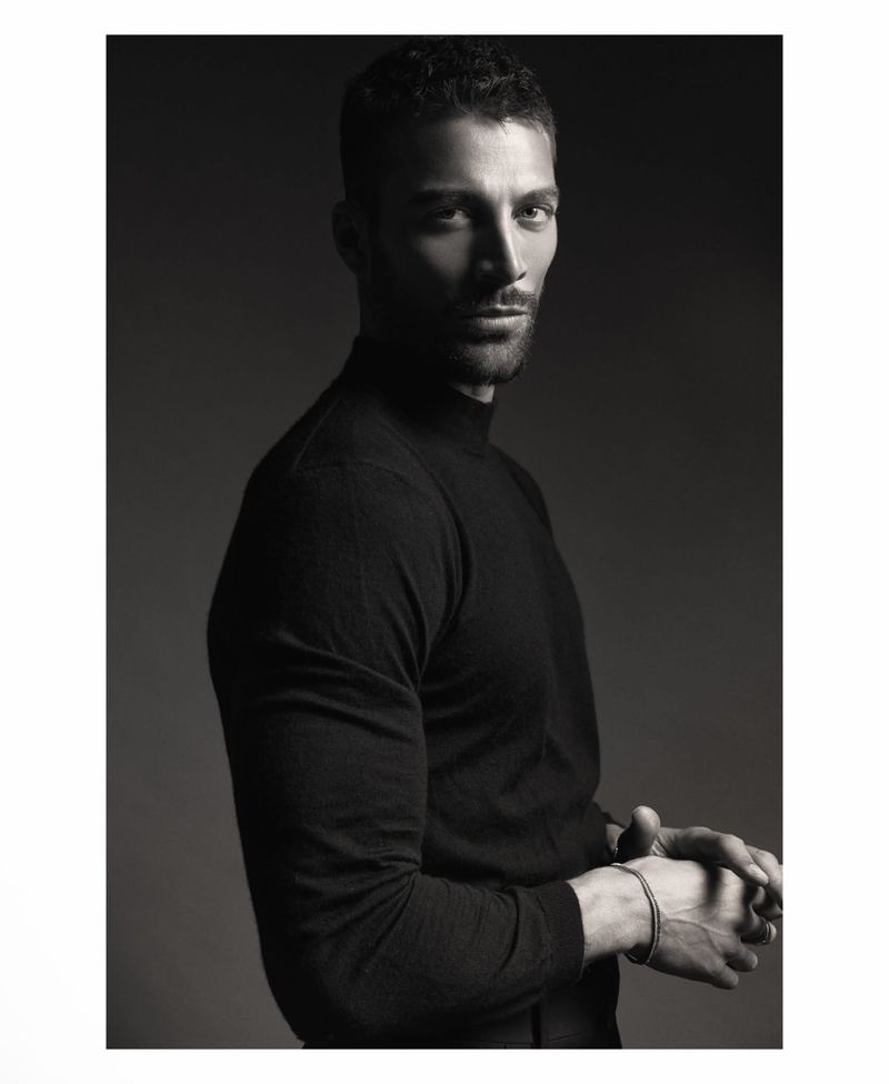 Monochrome studio portrait of model Simone Susinna wearing a black mock neck top with clasped hands against a dark background.