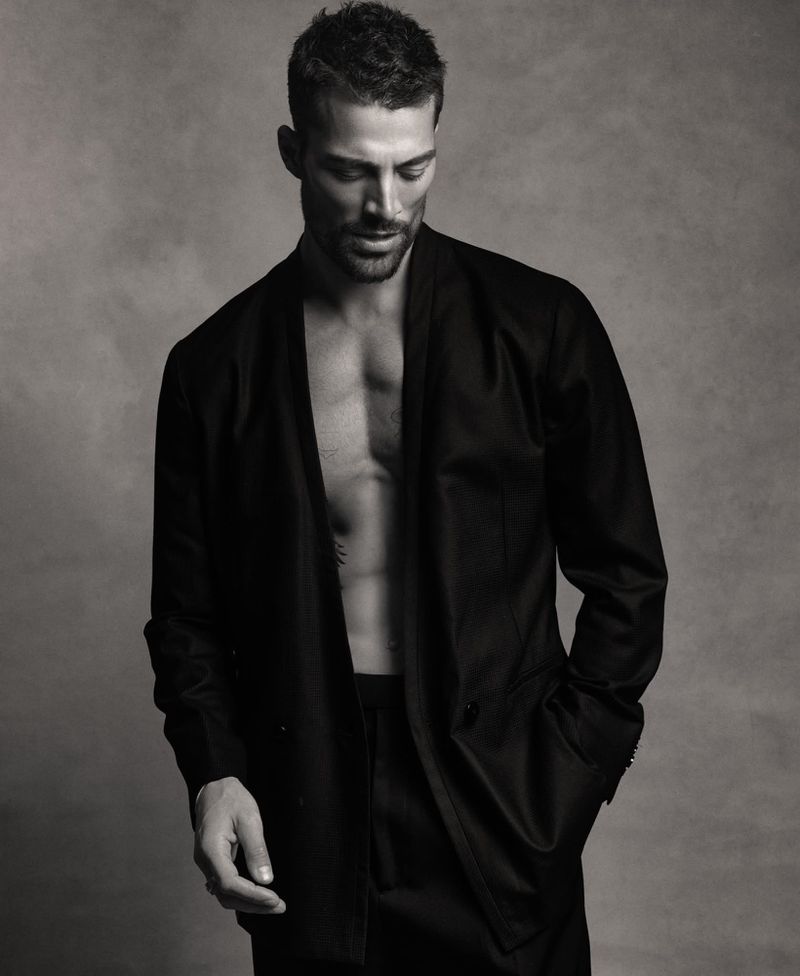 Monochrome portrait of Simone Susinna wearing an open black blazer with bare chest against a textured studio backdrop.