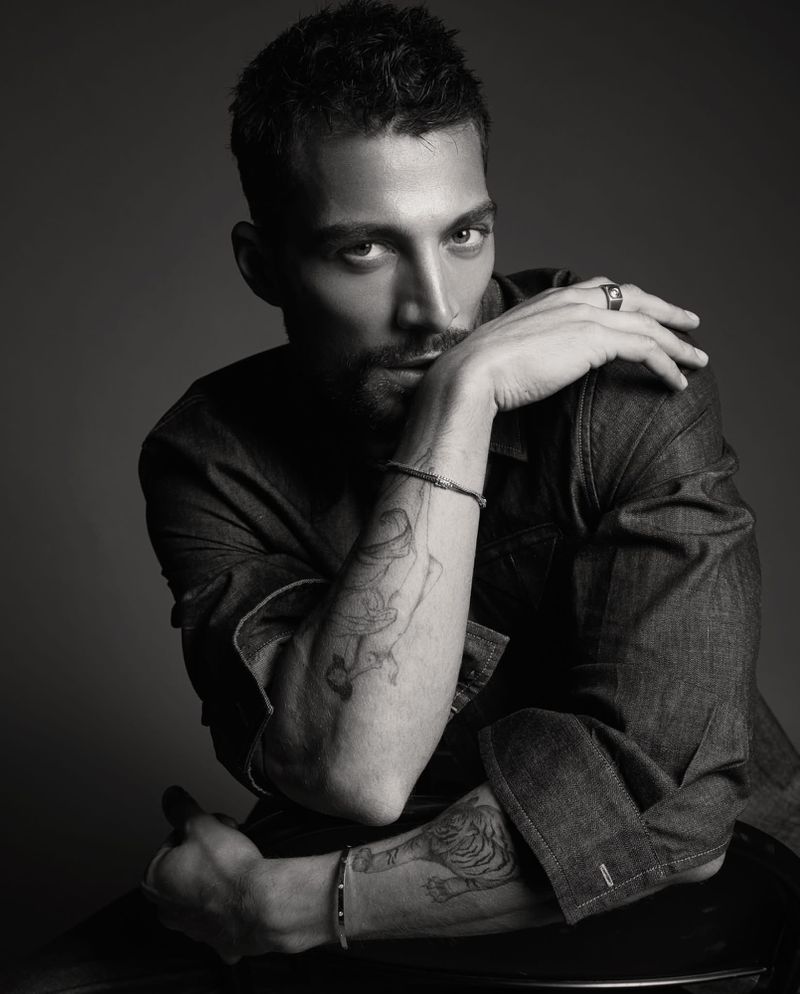 Black and white portrait of model Simone Susinna wearing a dark denim shirt with forearm tattoos against a studio backdrop.