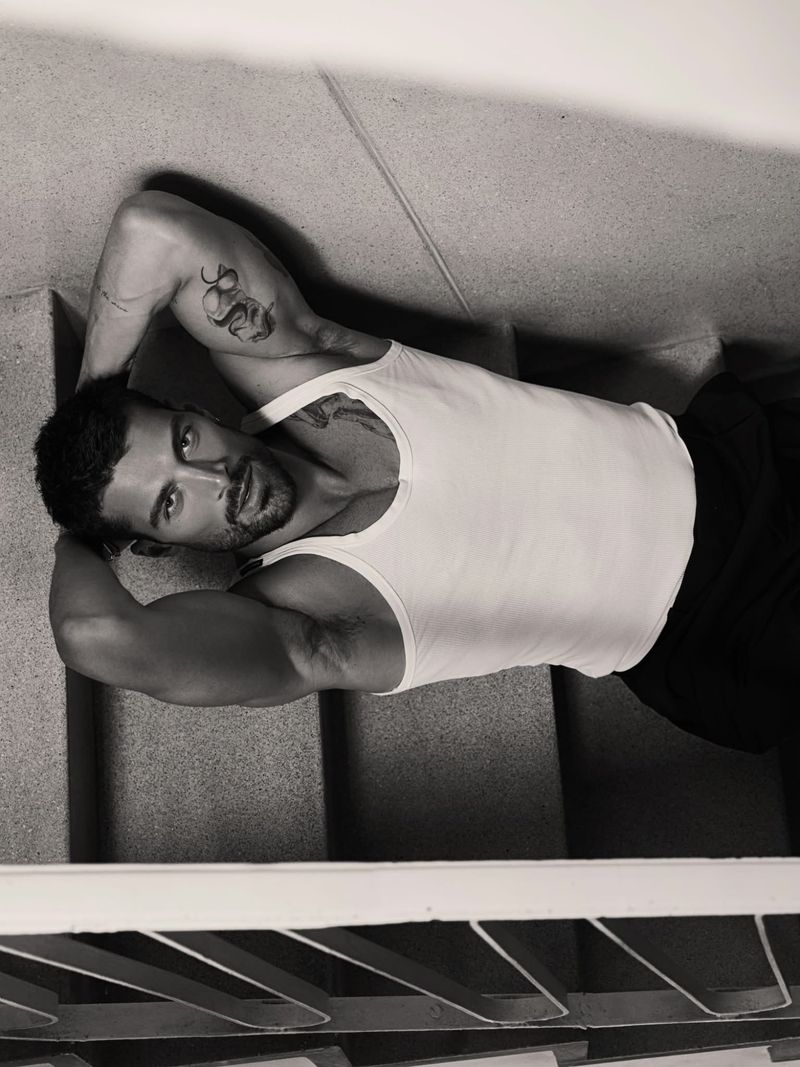 Model Simone Susinna wearing a white tank top lying on concrete stairs with hands behind his head in black and white.