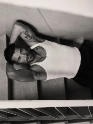 Model Simone Susinna wearing a white tank top lying on concrete stairs with hands behind his head in black and white.