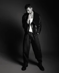 Simone Susinna posing in a black leather jacket, white tank top, and pleated dark trousers in a monochrome studio portrait.