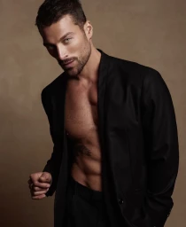 Simone Susinna wearing an open black blazer shirtless against a warm studio backdrop.