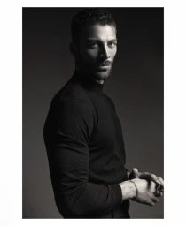 Model Simone Susinna posing in a black long-sleeved turtleneck top against a dark studio background in a low-key portrait.