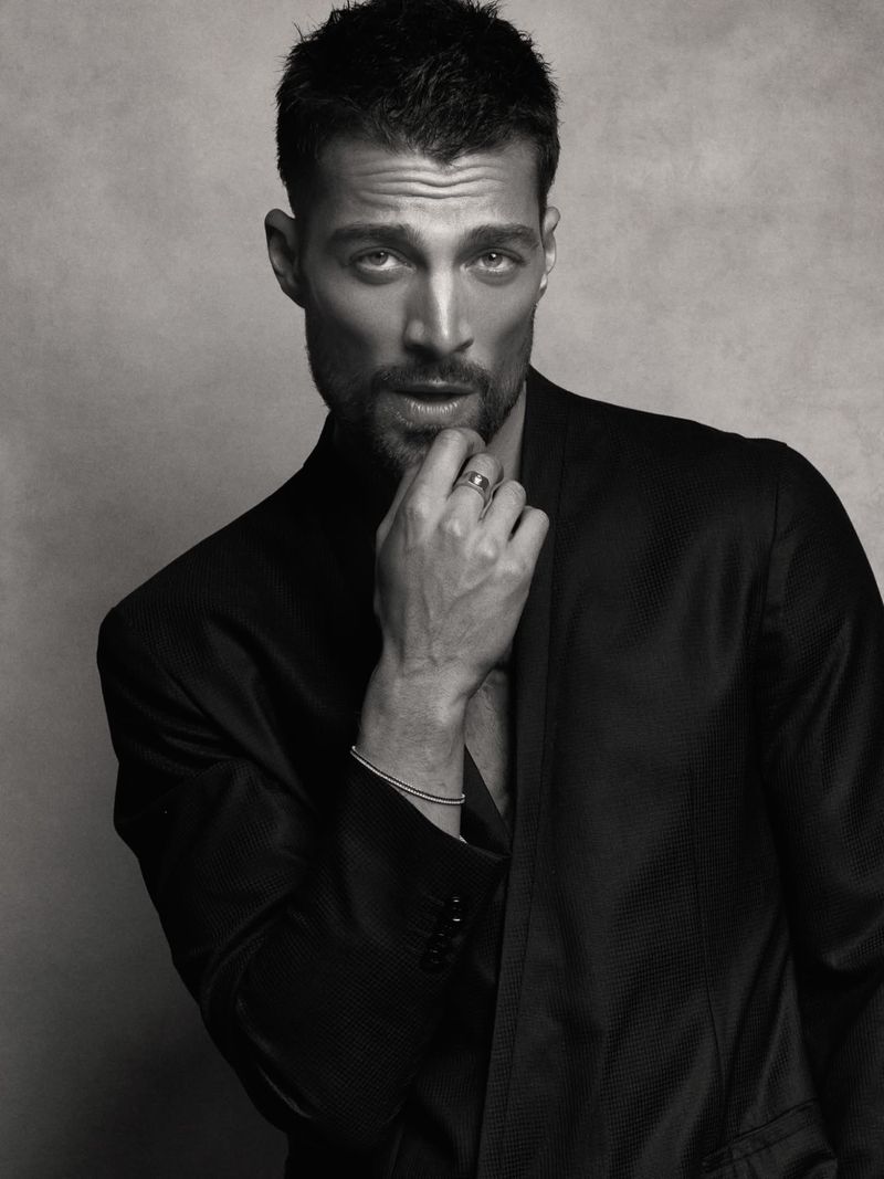 Monochrome portrait of model Simone Susinna wearing a black blazer with his hand touching his chin against a textured backdrop.