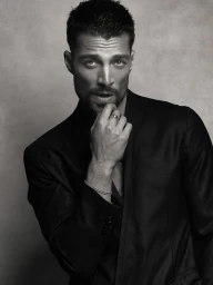 Monochrome portrait of model Simone Susinna wearing a black blazer with his hand touching his chin against a textured backdrop.