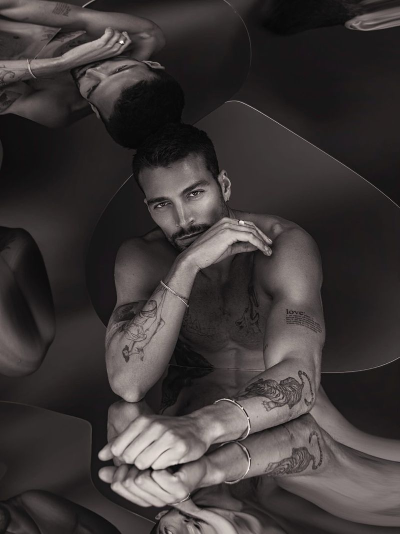 Monochrome portrait of tattooed model Simone Susinna posing shirtless over a reflective mirror surface wearing silver jewelry.