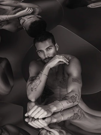 Monochrome portrait of tattooed model Simone Susinna posing shirtless over a reflective mirror surface wearing silver jewelry.
