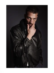 Model Simone Susinna wearing a black leather jacket with a knit collar posing with his hand on his chin in a studio portrait.