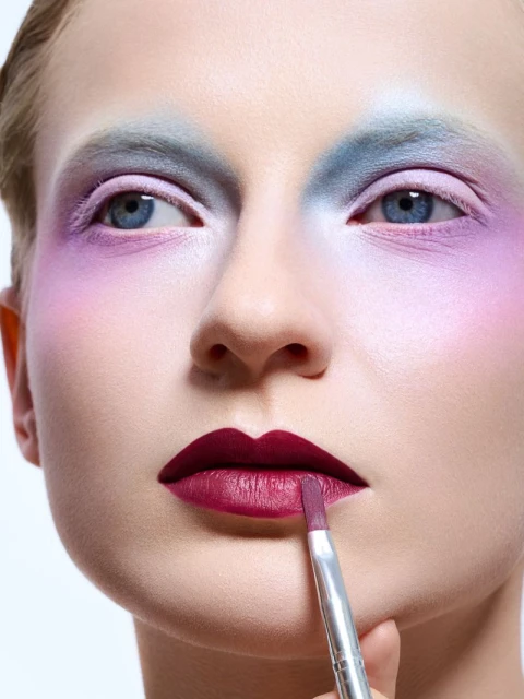 Close-up of fair-skinned woman with bold, colorful eyeshadow and dark lipstick, applying makeup with a brush against a white backdrop.