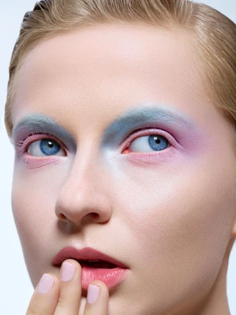 Close-up portrait of a fair-skinned adult female with blue eyes and stylized colorful makeup, posing indoors against a light background.