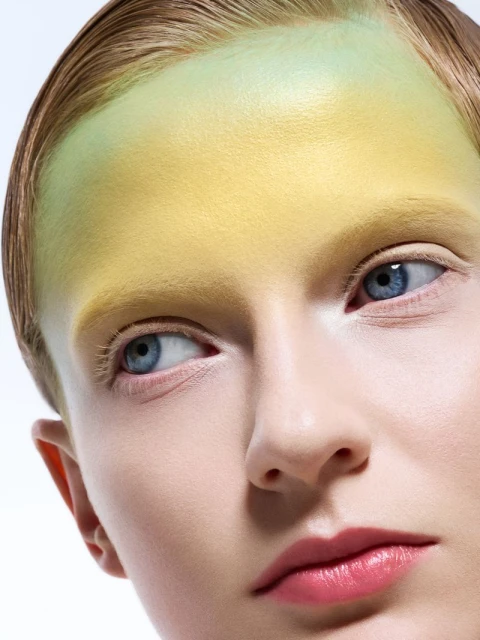 Close-up portrait of a woman with blue eyes and yellow and green makeup on her forehead against a white background in a beauty shot.