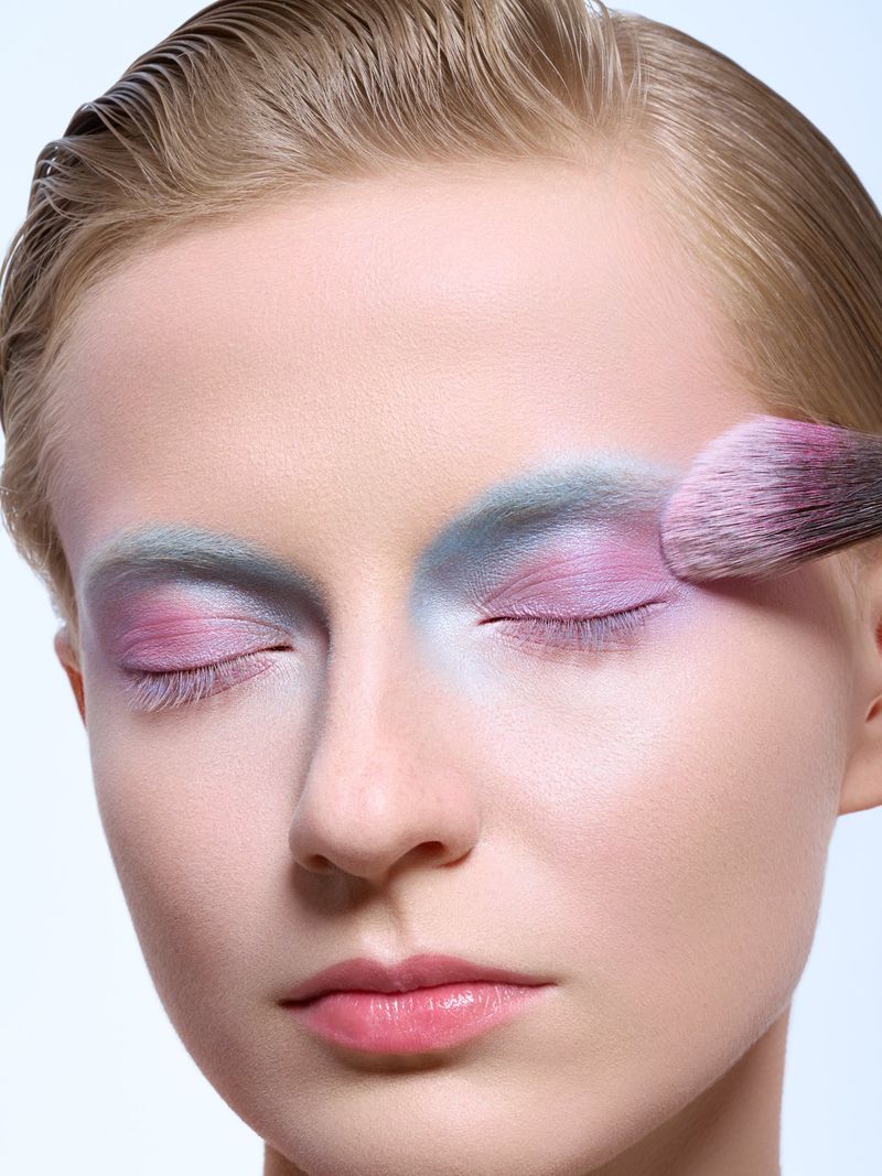 Close-up shot of a model receiving makeup application on her eyelids using a pink brush with fair skin and blonde hair in a beauty setting.