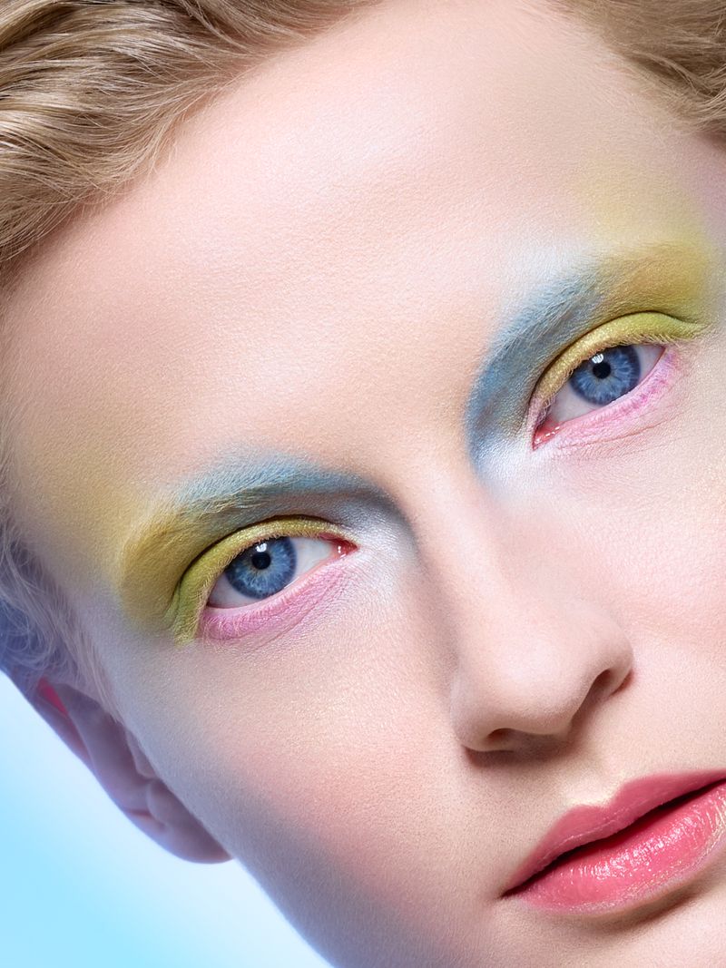 Close-up of a young woman's face with vibrant yellow, blue, and pink eyeshadow. A portrait highlighting her blue eyes and pink lips.