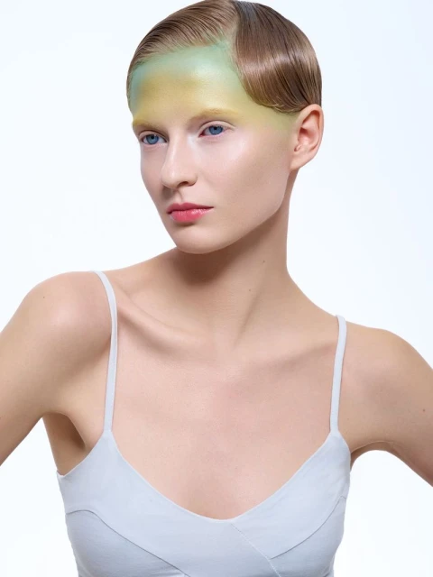 Close-up portrait of a female fashion model with slicked-back brunette hair and artistic green and yellow face makeup on a white backdrop.