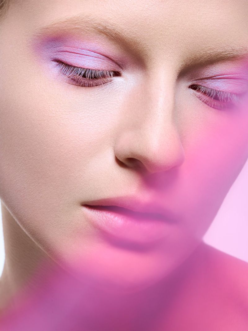 Close-up portrait of an adult female with closed eyes, wearing pink and purple eyeshadow and pink lipstick, with a light pink background.