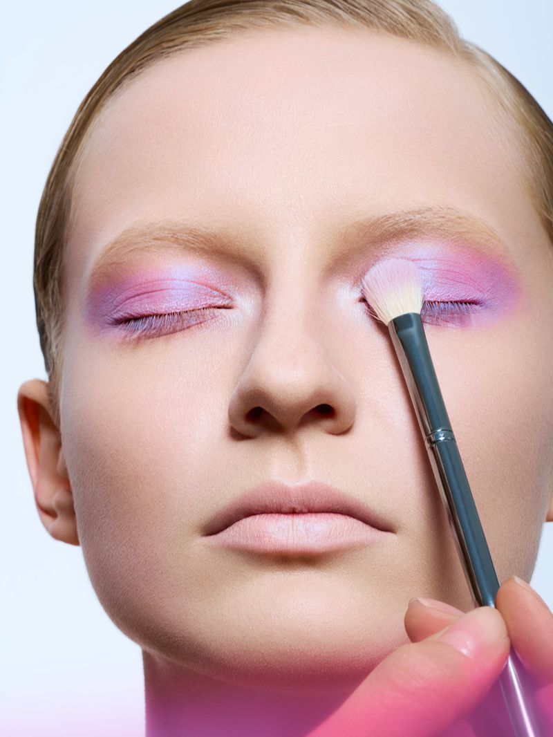 Close-up of a fair-skinned adult female with closed eyes having purple eyeshadow applied with a makeup brush in a beauty studio.