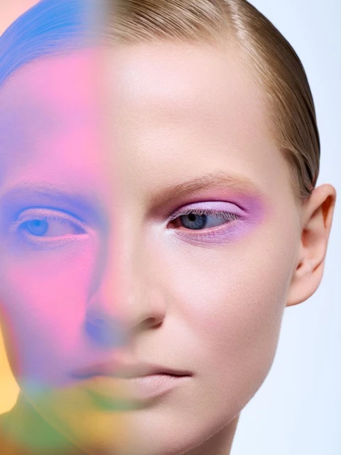 Close-up portrait of a fair-skinned adult woman with a slicked-back hairstyle and violet eyeshadow, partially lit with multicolor gel lighting.