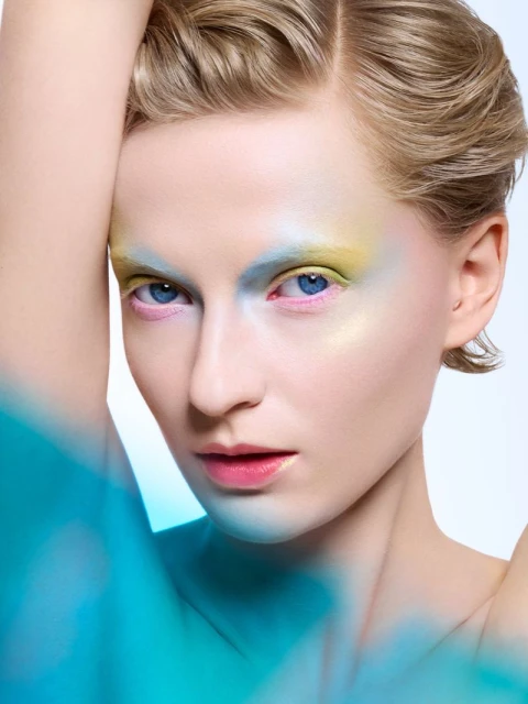 Close-up studio shot of a blond woman with colorful makeup. She poses with her arm raised behind her head in front of a bright background.