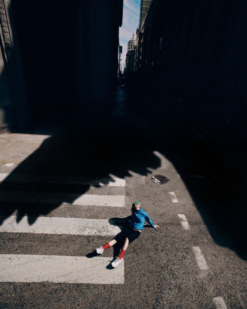 High-angle shot of a person sitting on an asphalt crosswalk wearing a blue long-sleeve top, dark shorts, red socks, and white sneakers.