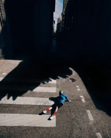High-angle shot of a person sitting on an asphalt crosswalk wearing a blue long-sleeve top, dark shorts, red socks, and white sneakers.