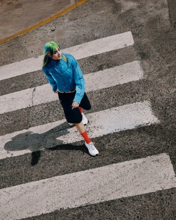 High-angle shot of a woman with green hair wearing a blue jacket, black shorts, and white sneakers walking on a crosswalk.