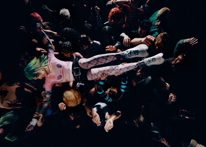 Top-down view of a woman with green hair crowd surfing over hands wearing white athletic running shoes and patterned tights.