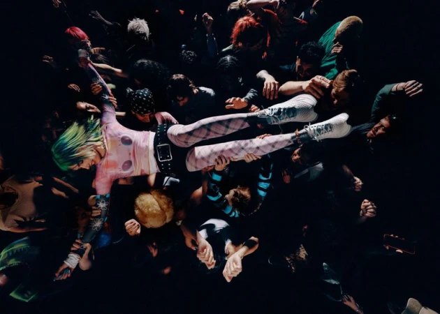 Top-down view of a woman with green hair crowd surfing over hands wearing white athletic running shoes and patterned tights.