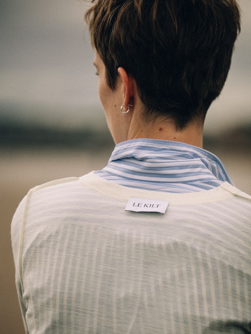 Back view of a person with short dark hair wearing a white top with a Le Kilt label over a blue striped collar.
