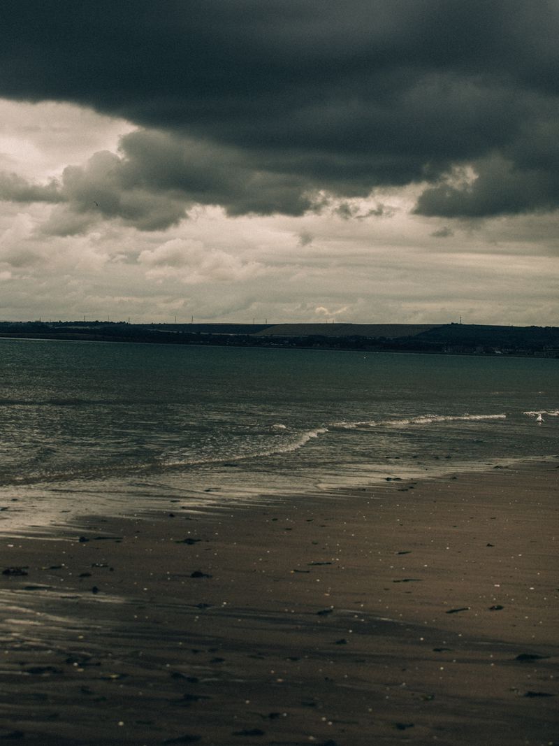 Moody coastal view of wet sand, gentle ocean waves, and heavy dark storm clouds over a distant coastline.