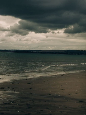 Moody coastal view of wet sand, gentle ocean waves, and heavy dark storm clouds over a distant coastline.