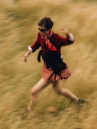 Model wearing a red sweater, neck scarf, and pleated kilt running across a dry grass field captured with motion blur.