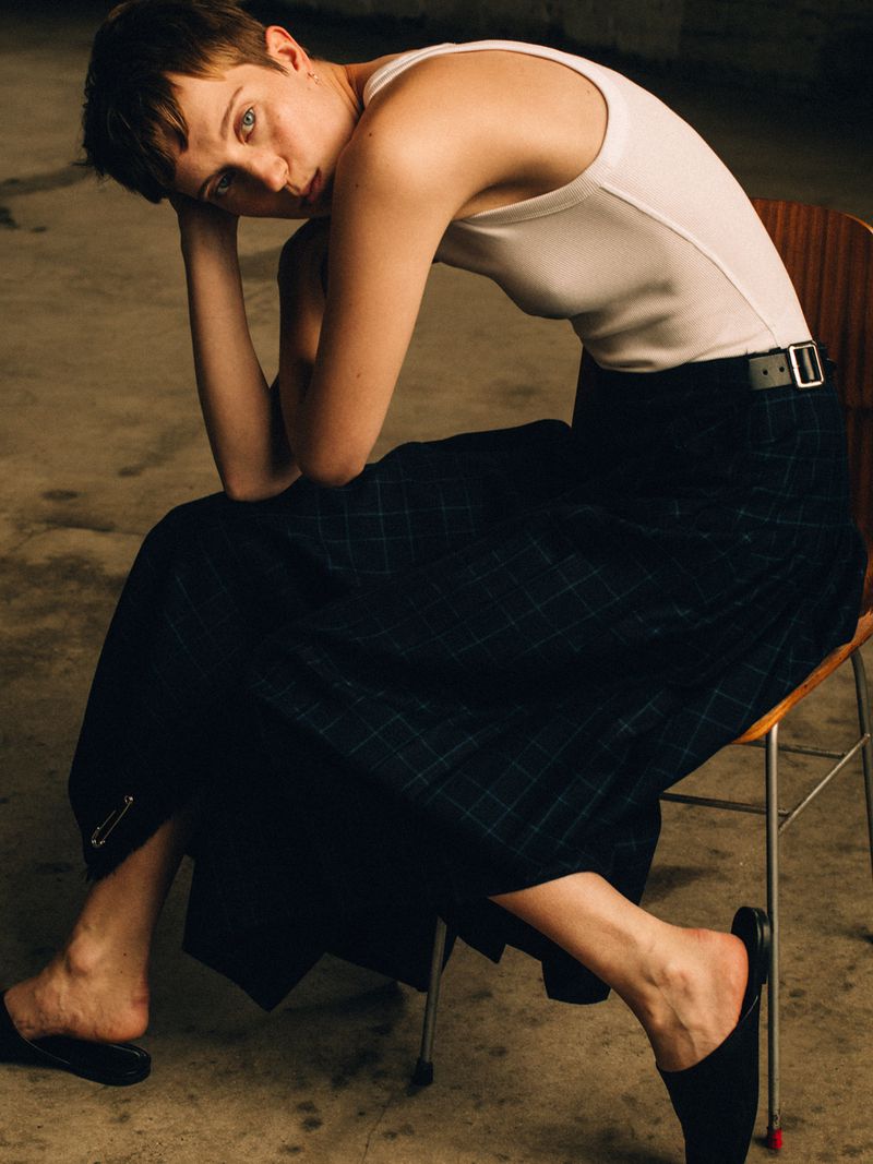 Model with short hair wearing a white tank top and dark pleated tartan skirt seated on a wooden chair.