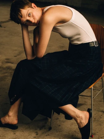 Model with short hair wearing a white tank top and dark pleated tartan skirt seated on a wooden chair.