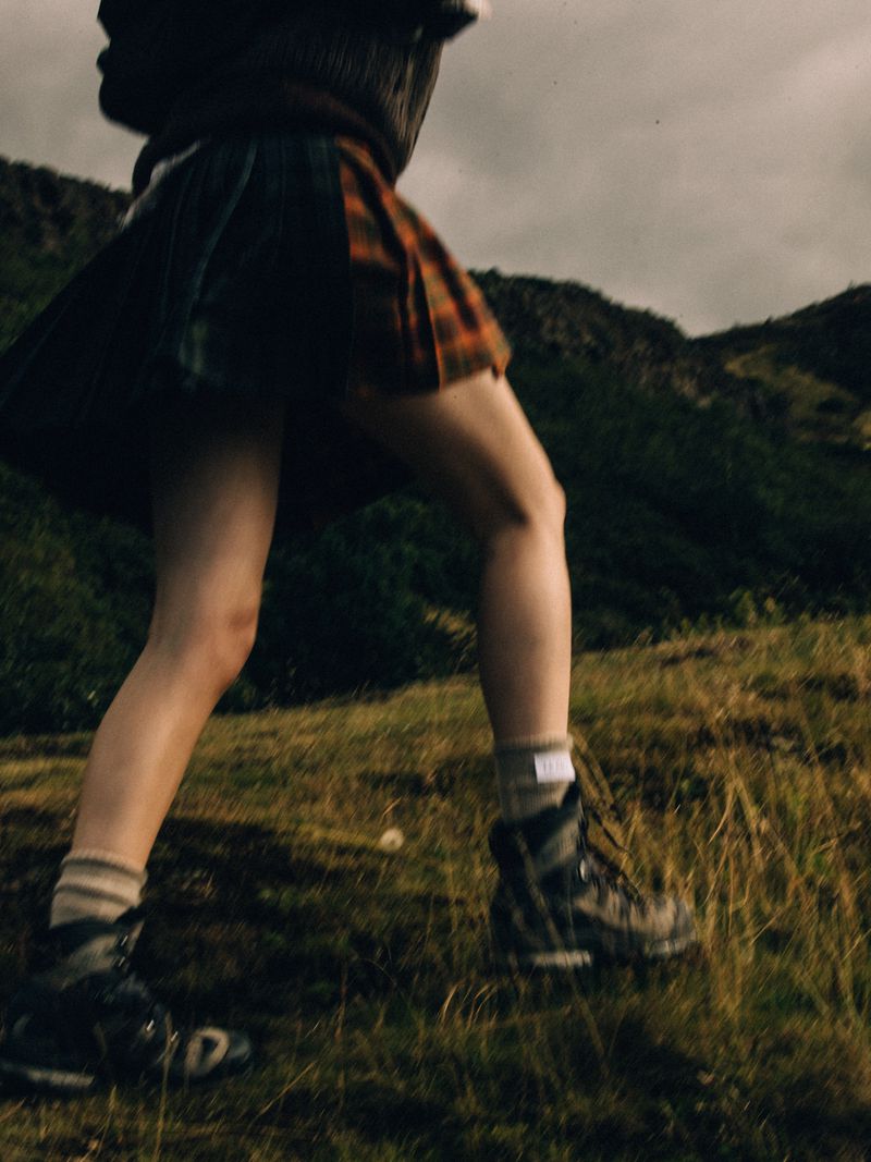 A low-angle view of a person wearing a pleated tartan kilt and hiking boots while walking up a grassy hill under an overcast sky.