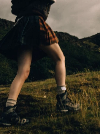 A low-angle view of a person wearing a pleated tartan kilt and hiking boots while walking up a grassy hill under an overcast sky.