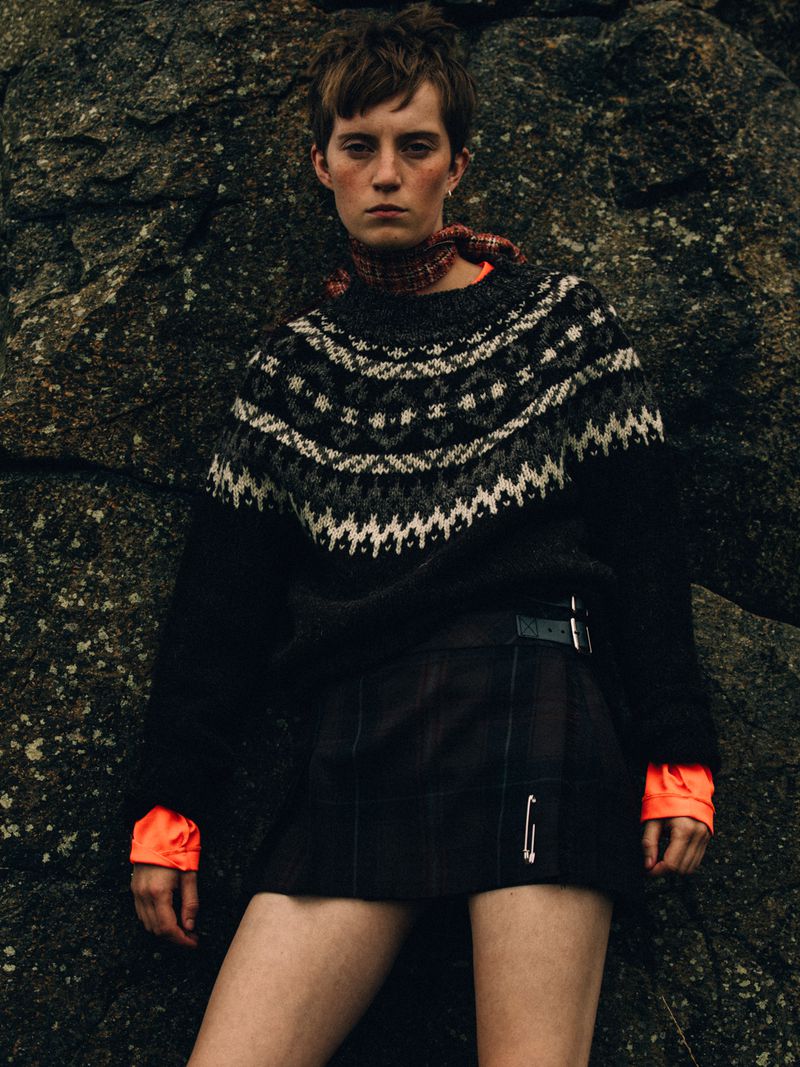 Model in a black Fair Isle knit sweater and pleated kilt mini skirt standing against a dark mossy rock surface.