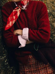 Cropped view of a model wearing a red knit sweater, striped cuffs, and a patterned red neckerchief leaning against a stone surface.