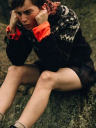 Model sitting on a lichen-covered rock wearing a black patterned knit sweater with neon orange cuffs and dark shorts.