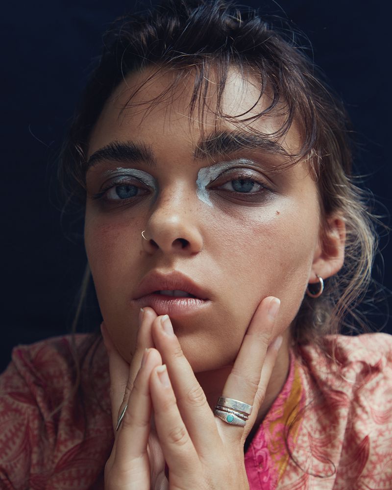 Close-up studio shot of a young adult female model with creative white eye makeup and rings, posing with her hands near her face.