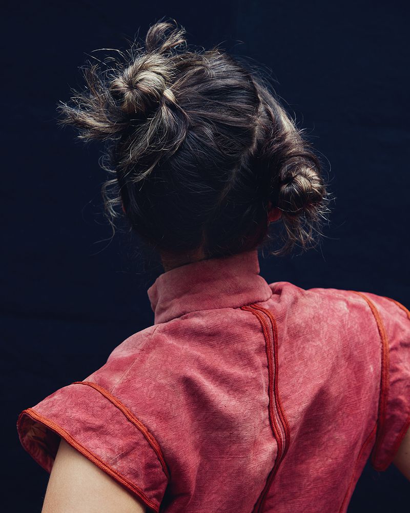 Woman with braided updo wearing a coral-colored high-necked top against a dark blue background, shot from the back showcasing hairstyle.